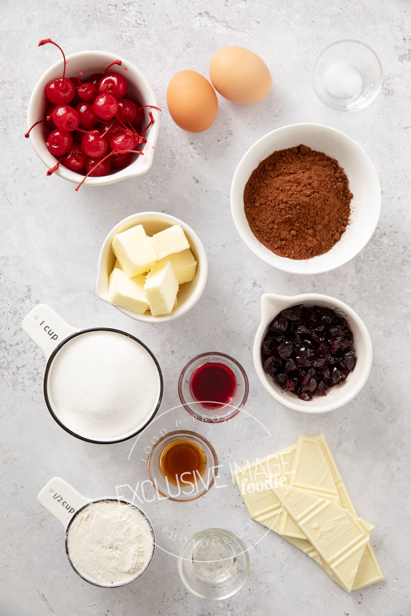 Ingredients for 2 bite cherry brownies recipe including cherries, eggs, cocoa powder, butter, sugar, and vanilla extract on a gray surface.