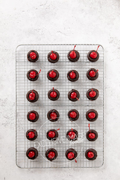 Chocolate mini brownies with red cherries on a cooling rack against a light gray background