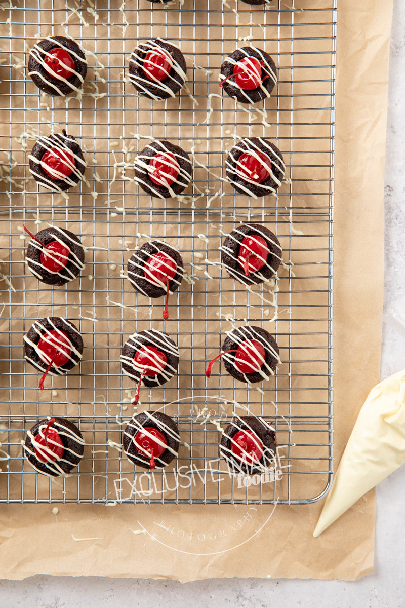 Chocolate 2 bite brownies with red cherries on a cooling rack and a piping bag filled with white chocolate nearby.