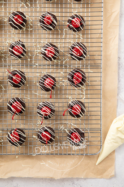 Chocolate 2 bite brownies with red cherries on a cooling rack and a piping bag filled with white chocolate nearby.