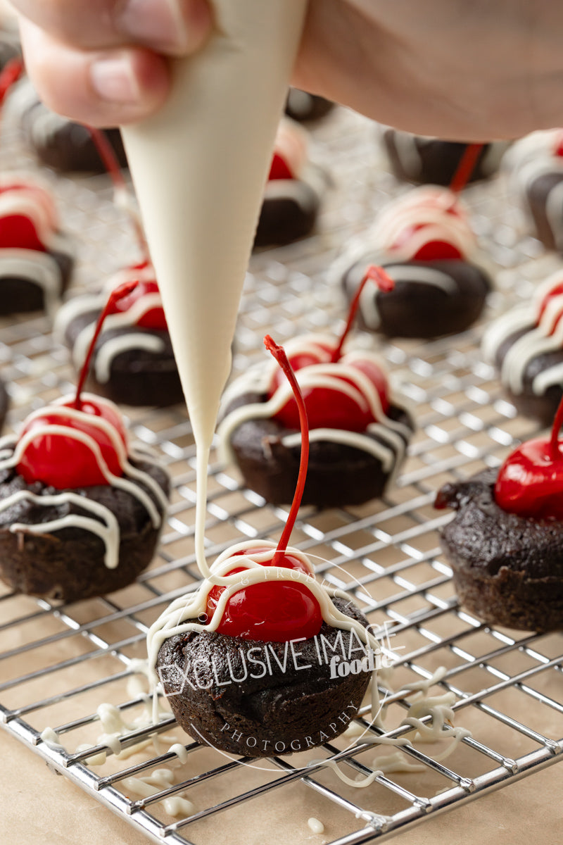 Chocolate brownie bites with cherries being decorated with white chocolate on a cooling rack.
