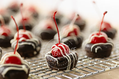 Chocolate 2 bite brownies with white drizzle and red cherries on a cooling rack.