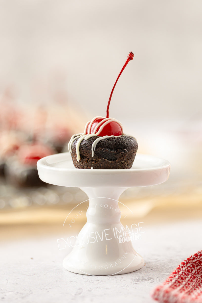 Small chocolate brownie with white icing and a cherry on top, placed on a white stand against a blurred background.