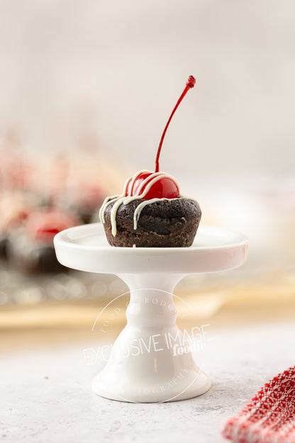 Small chocolate brownie with white icing and a cherry on top, placed on a white stand against a blurred background.