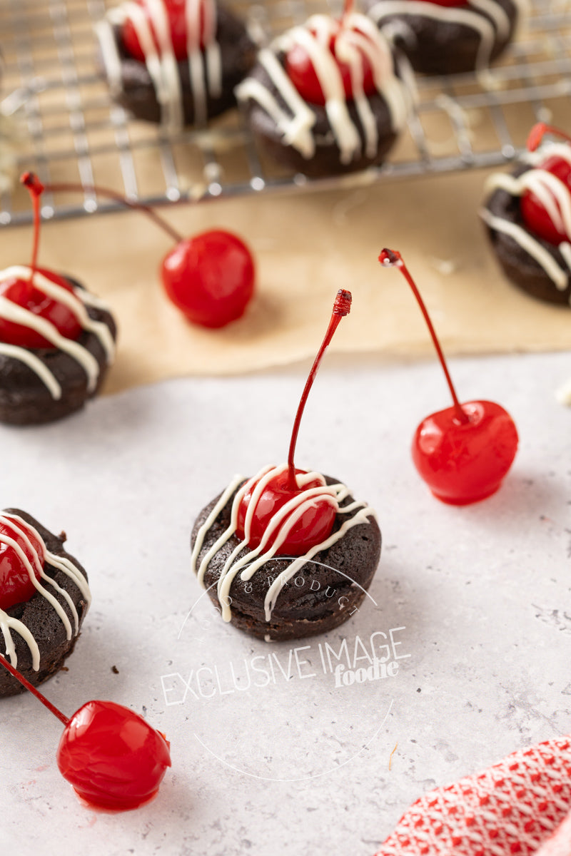 2 bite brownie treats with cherries on a light surface, with a cooling rack in the background.
