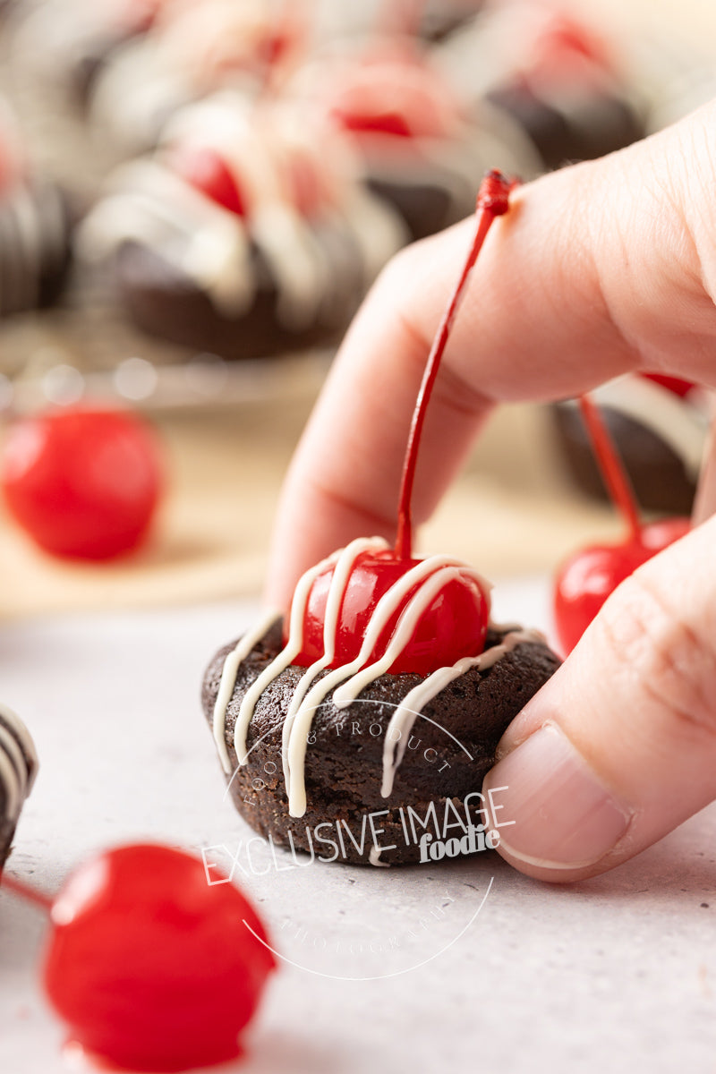 Chocolate brownie bite with a cherry on top held by a hand, with more brownies and cherries in the background.
