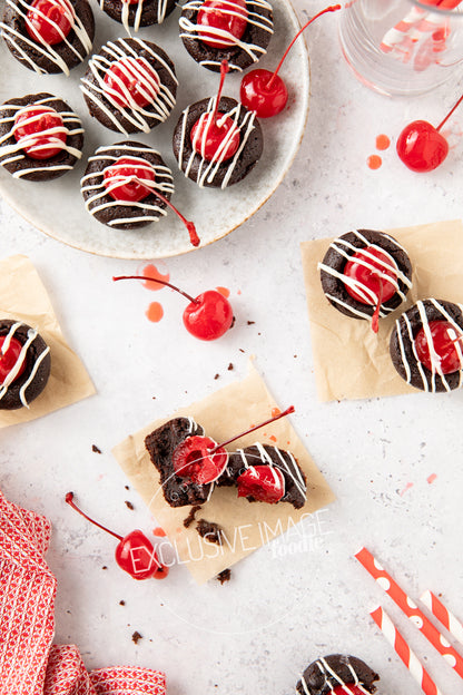 Chocolate 2 bite brownies with cherry toppings on a white surface with red straws and a red napkin.