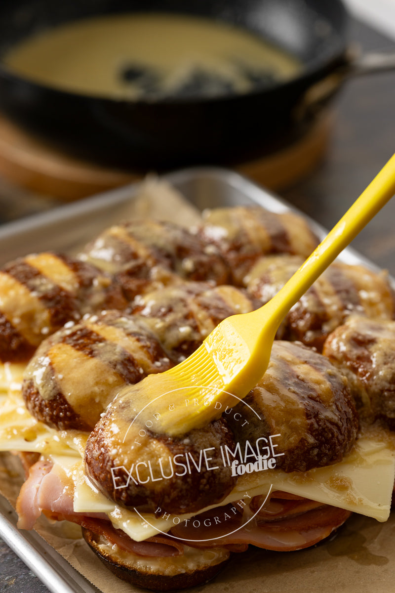 Tray of sandwiches with cheese and ham, on pretzel bun sliders, on a tray with a yellow spatula spreading butter.