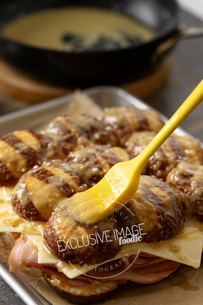 Tray of sandwiches with cheese and ham, on pretzel bun sliders, on a tray with a yellow spatula spreading butter.