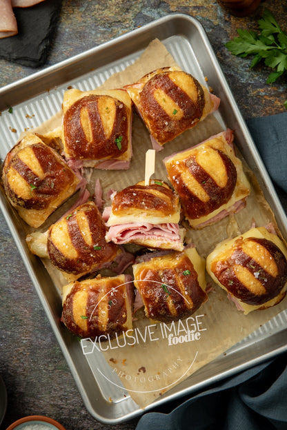 Baked ham and cheese sliders on a baking tray with linen and ingredients around.