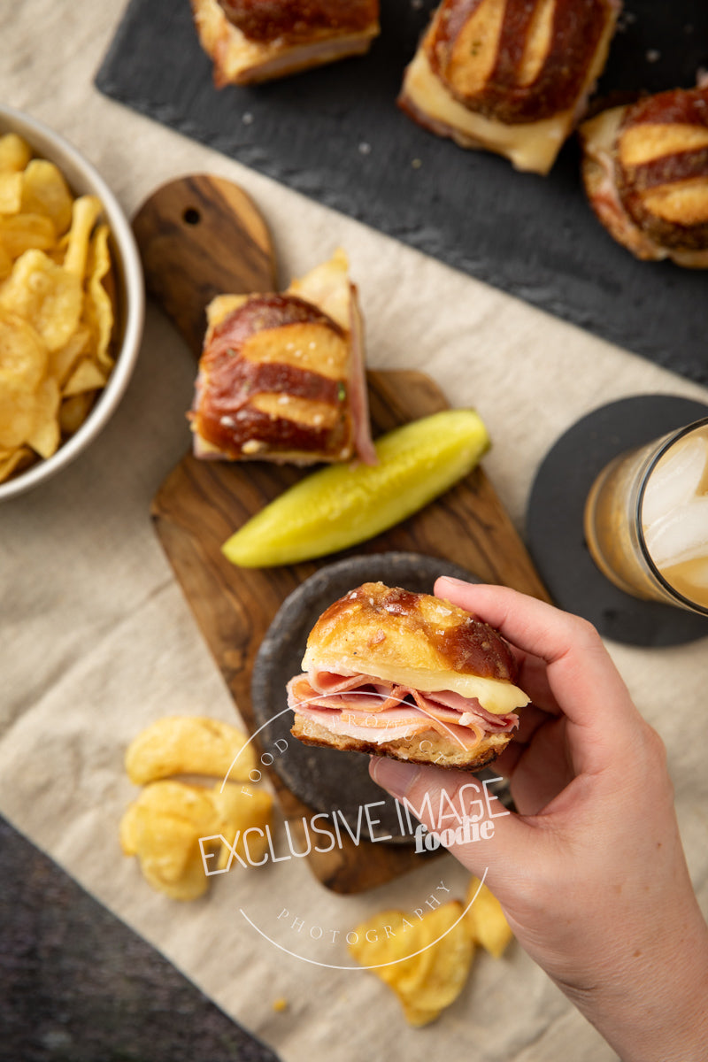 Hand holding a ham and cheese slider with a bowl of chips and a drink on a wooden board.