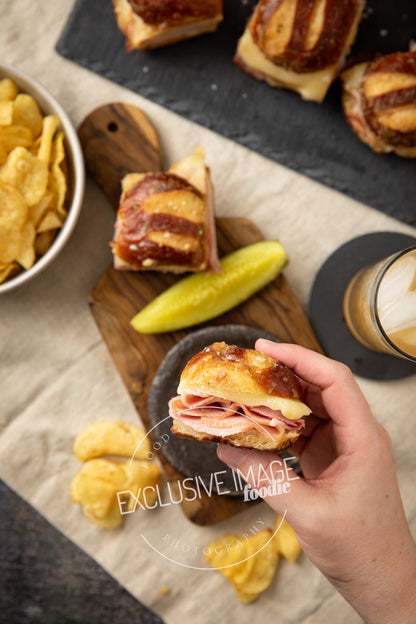 Hand holding a ham and cheese slider with a bowl of chips and a drink on a wooden board.