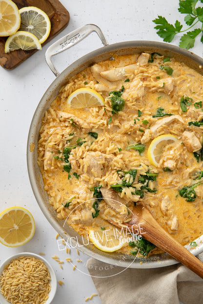 Lemon chicken and rice dish in a pan with wooden spoon, surrounded by lemons and herbs.