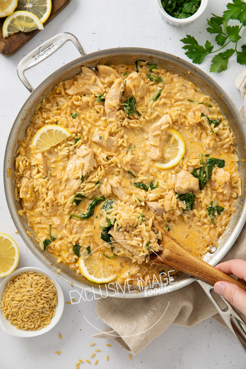 Skillet of creamy lemon chicken and rice with a wooden spoon, surrounded by lemon slices and herbs on a light background.