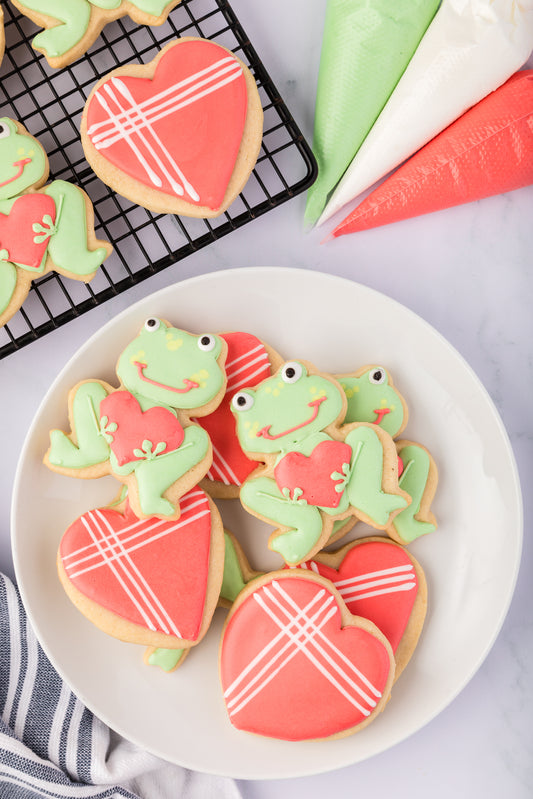 Decorative cookies shaped like frogs and hearts on a plate with a cooling rack in the background.