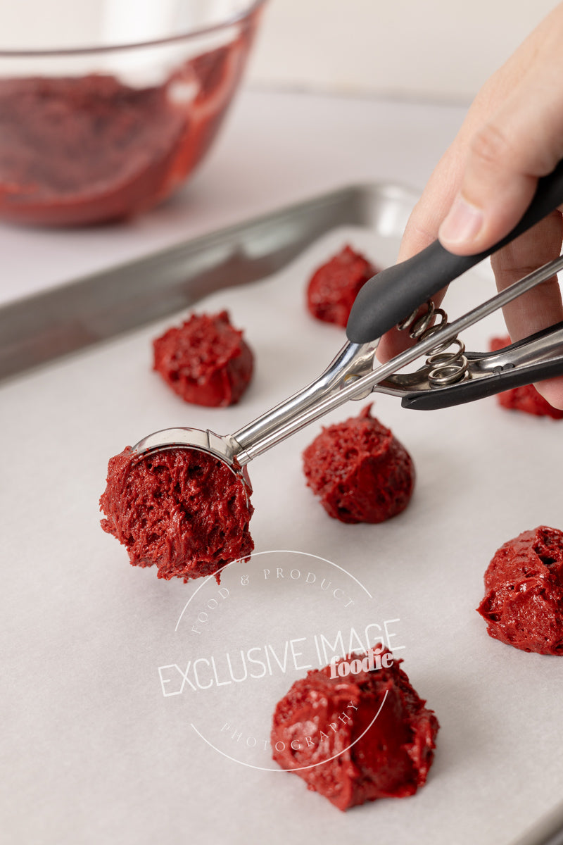 Red velvet cookie dough being scooped onto a baking sheet with a cookie scoop.